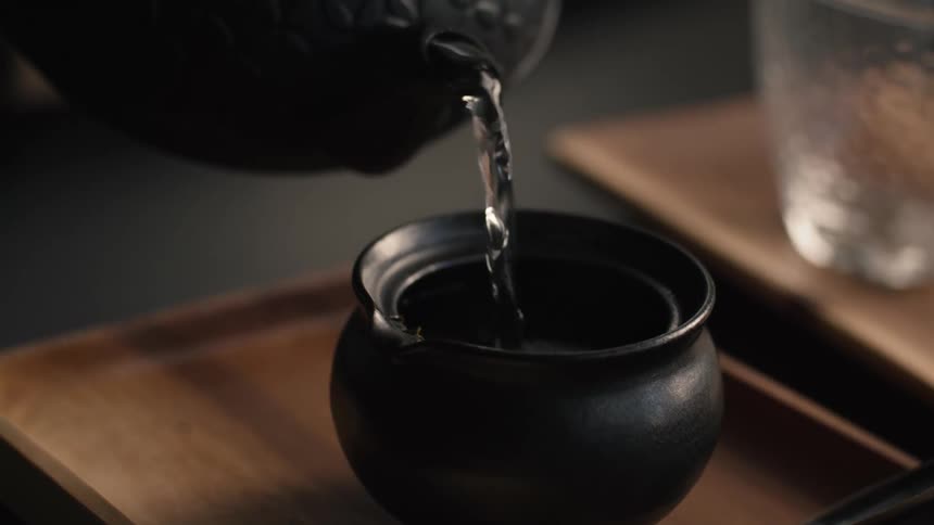 Hot water pours gracefully into a dark ceramic cup set on a wooden tray evoking a calm traditional tea moment