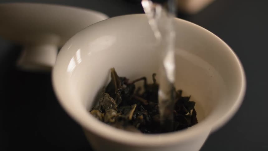A very close view shows hot water being poured from above into a small white cup containing dark green loose leaf tea