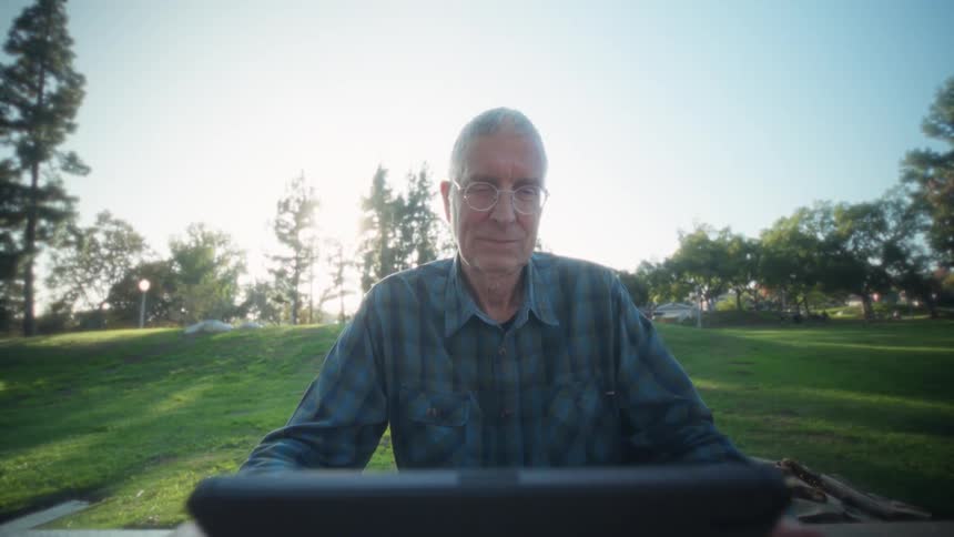 Elderly man sitting down at a park using a tablet computer over a wooden table while smiling outdoors during dusk or dawn