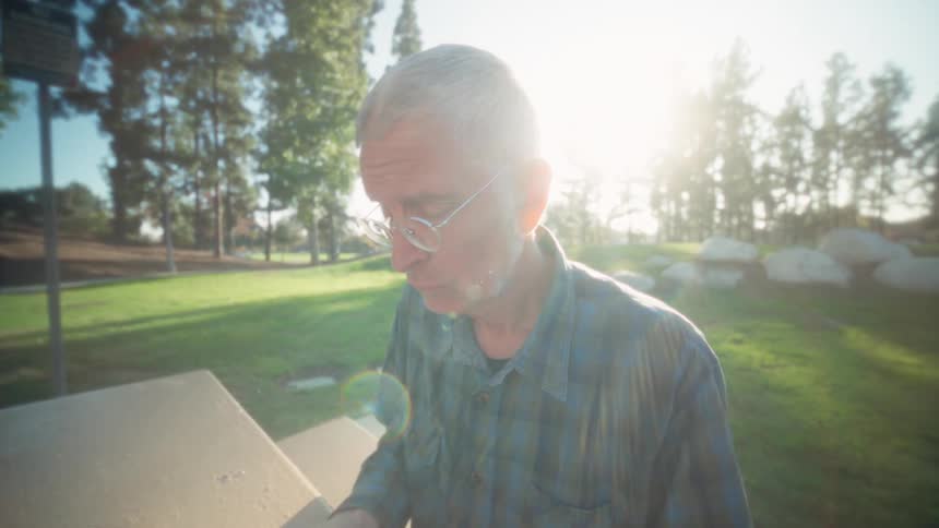 A close view of an older man with glasses wearing a plaid shirt in a sunny park setting looking down and concentrating on a task