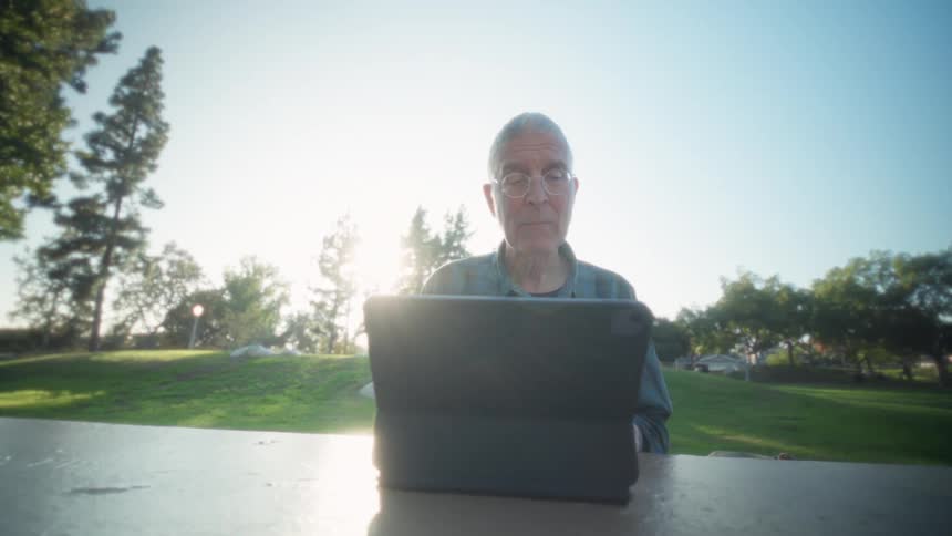 An elderly man wearing glasses types on a computer keyboard while sitting on a bench in a public park on a sunny day