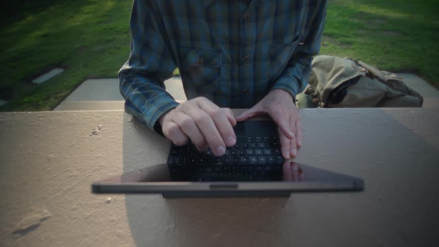 An elderly man with glasses wearing a plaid shirt sits at a concrete table in a sunny park and opens a tablet attached to a keyboard case