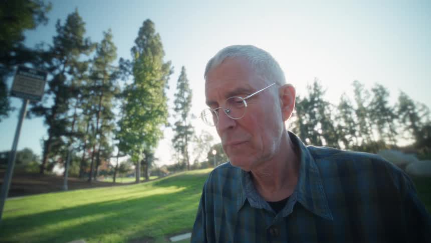 Elderly man wearing a plaid shirt and glasses sitting down on a park using a laptop on a table outdoors during dusk or dawn