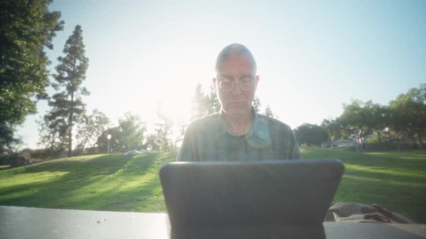 Man using a tablet while seated at an outdoor table in a park during daylight