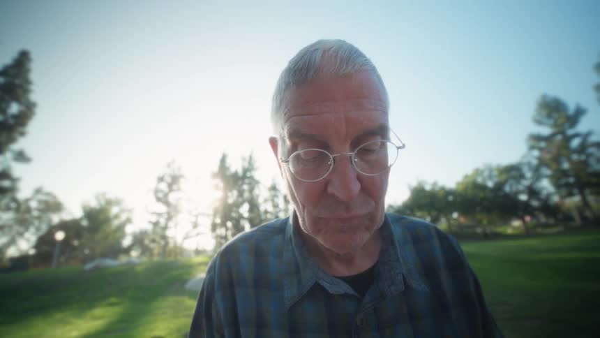 The older man is sitting at a table in the park using his laptop