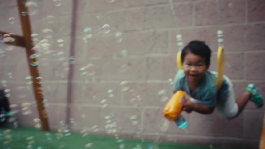 Two young child on a yellow swing in a backyard a boy wearing a blue shirt and a girl wearing a green dress laughs while enthusiastically holding and using an orange bubble machine that fills the frame with floating bubbles