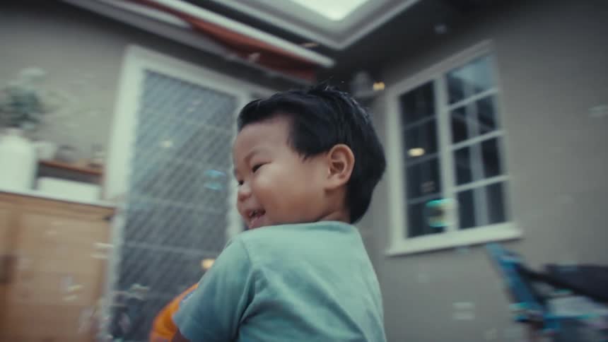 An excited boy in a light blue shirt runs on a paved backyard patio while shooting bubbles from an orange and yellow toy gun on a bright day