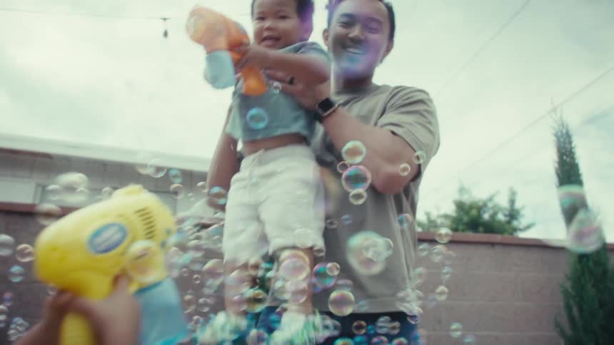 A father holds his young son as they and an older sister joyfully play with bubble guns in a backyard while the mom watches surrounded by many floating bubbles with the whole family smiling