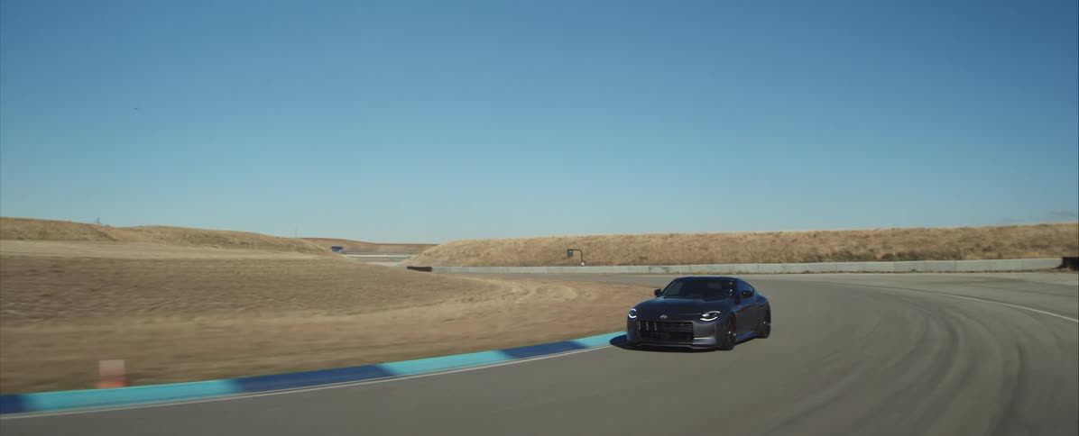 A black compact car navigates a curve on a paved race track surrounded by dry rolling hills