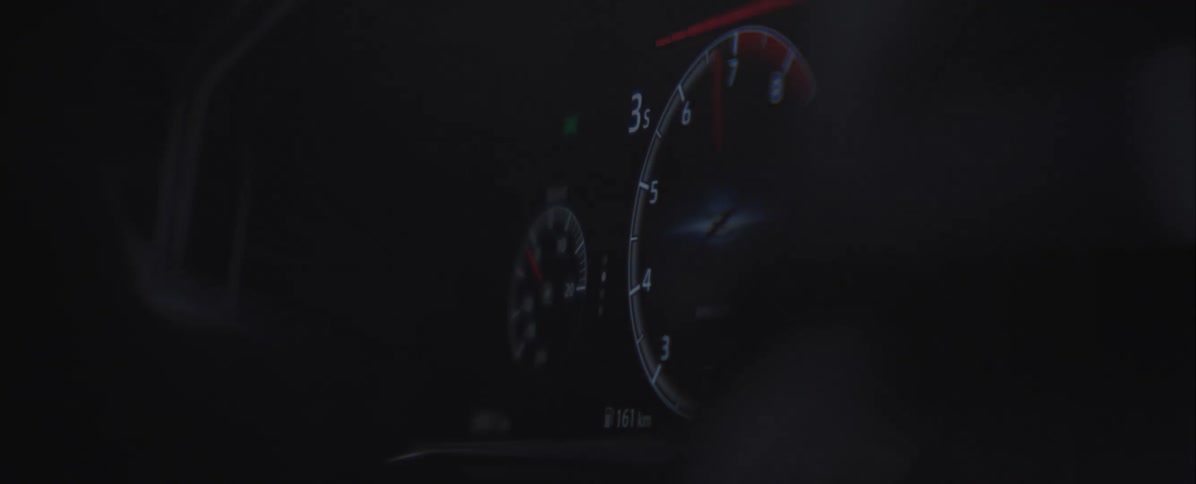 The illuminated needle of the tachometer glows brightly in the dark cabin hovering steadily near the numbers as blurred reflections of passing lights flicker across the shadowed foreground