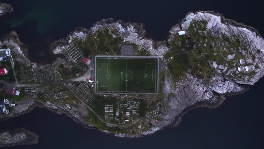 Soccer field on rocky island surrounded by dark water with buildings and fishing racks nearby