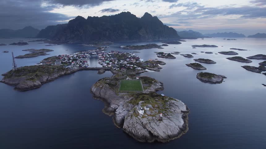 A small denselybuilt coastal village situated on a group of rocky islands featuring a prominent green football field with massive jagged mountains in the background under a dramatic cloudy sky