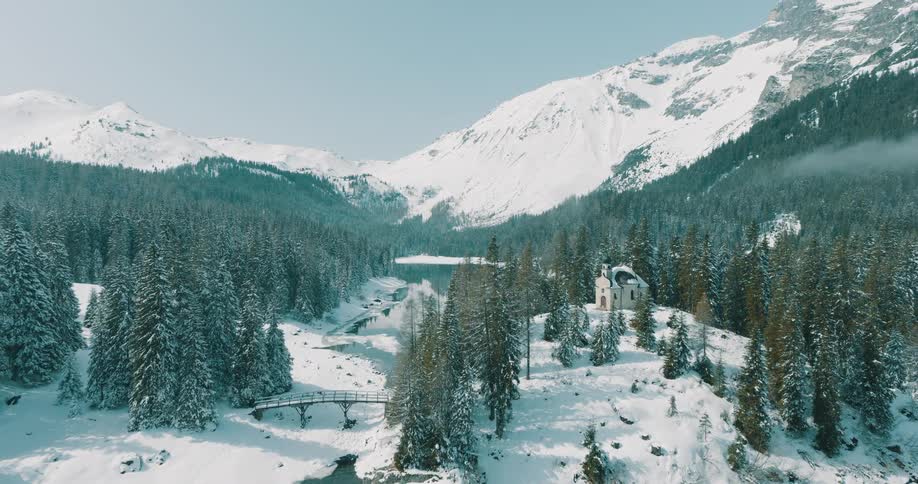 Rising above the icy waters of an alpine lake the camera reveals a picturesque chapel and bridge surrounded by a dense snowcovered pine forest and towering mountains