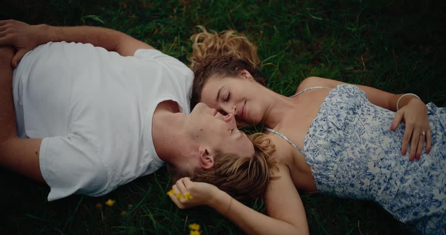 The video shows an overhead closeup shot of a young couple lying together on their backs in green grassthey are resting their heads next to each other and smilinggazing affectionately as the woman gently touches the mans face