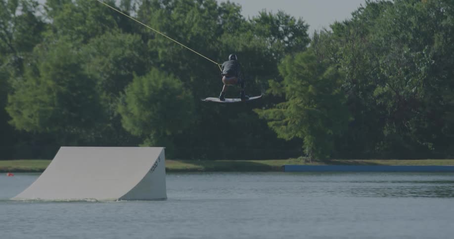 A man performs a trick on a wakeboard launching high in the air off a white ramp in a lake before landing with a splash surrounded by green trees