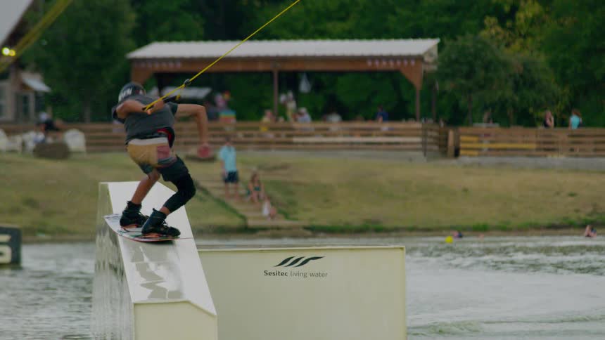 Man flies over a ramp and twists on his wakeboard as he holds a rope as people watch on the coast