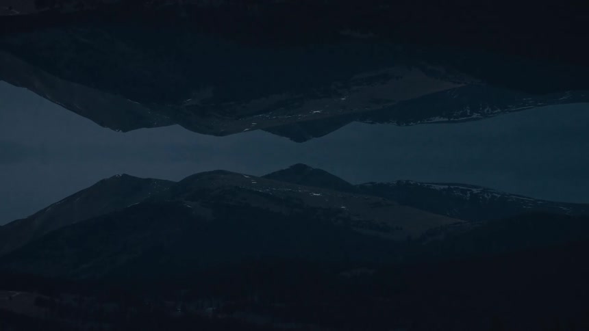 A dark symmetrical landscape of mountain silhouettes mirrors itself across a central horizon creating a calm abstract composition in deep blue tones