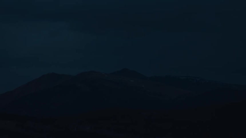 A dark somber shot of layered mountain peaks at twilight or night with a dark blue sky above