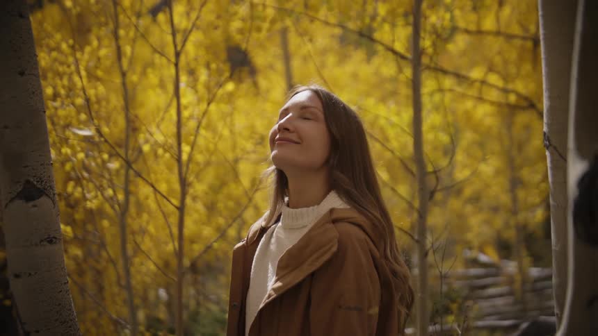 A woman stands peacefully among slender trees in a sunlit autumn forest her eyes closed and face tilted upward with a gentle smile as golden leaves glow around her in the warm afternoon light