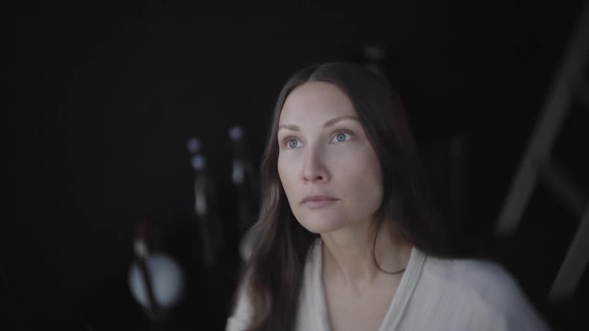 A thoughtful woman gazing slightly upward her calm expression emphasized by shallow focus and a dark unobtrusive background with bottles on a table