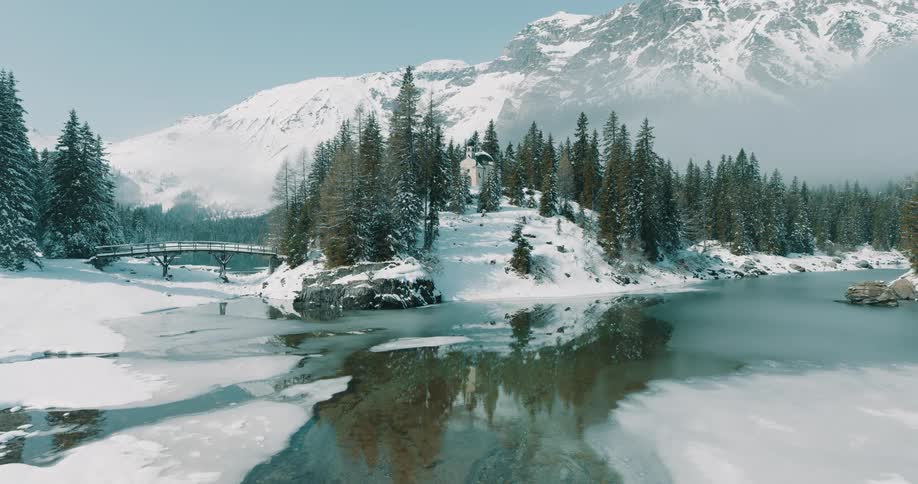 A snowy alpine landscape featuring a partially frozen lake pine trees a small wooden bridge and towering snow covered mountains under a bright sky