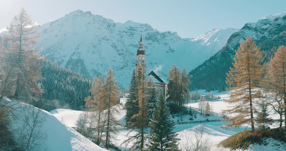 A sunlit church stands in a snowy mountain valley among evergreen trees