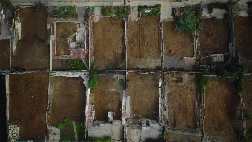 A top down drone shot slowly ascends revealing a grid of rectangular dirt plots and concrete foundation remnants on a large vacant lot showing property lines