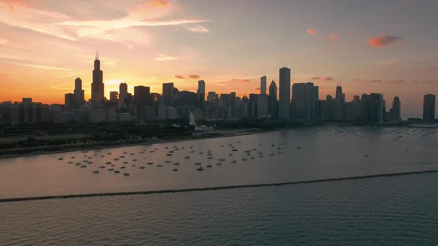 An aerial perspective glides across calm waters toward a sprawling city skyline where towering skyscrapers stand silhouetted against a vibrant orange sunset that illuminates a marina full of anchored boats