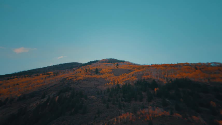 The view dramatically zooms out from a rugged mountain summit revealing steep slopes blanketed in a stunning contrast of vibrant orange autumn leaves and deep green evergreens