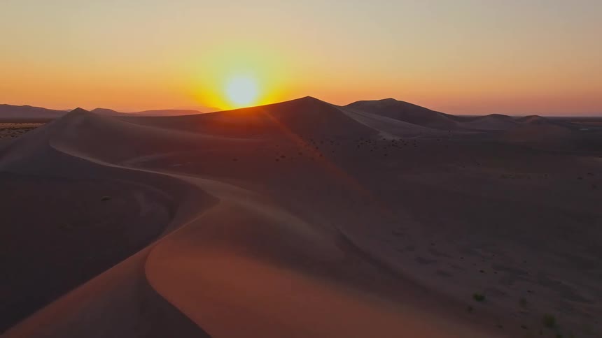 The video is an aerial view of vast sand dunes in a desert landscape as the sun sets the sky glows with warm orange and yellow light which highlights the curves and shadows of the massive dark dunes below