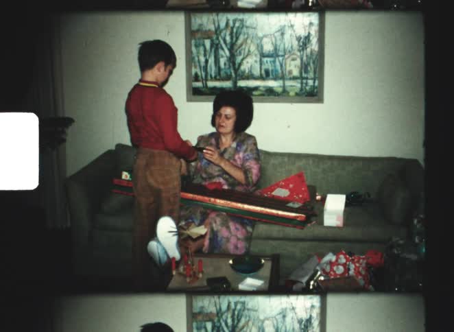 A woman sitting on a green sofa wrapping a gift with patterned paper as a young boy in a red shirt stands next to her leaning in all set in a cozy room with framed paintings on the wall