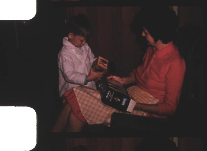 This is a short silent video clip of a woman and a young child indoors the child wearing a white jacket is presenting a small wrapped box to the seated woman who smiles while holding another larger box