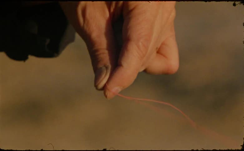 The video is a closeup shot of a persons hand holding a thin pale pink piece of string or thread the fingers gently pinch and release the string against a blurry warmtoned background of earth or sand