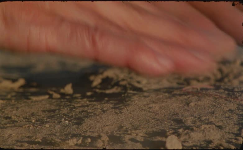 A persons arm is seen playing with beach sand at midday spreading the grains across a red thread lying on the ground