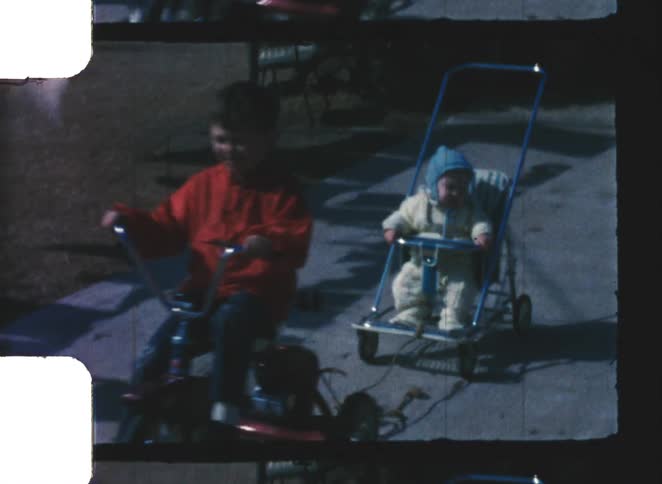 A boy is riding a tricycle and pulling a baby bundled in a winter outfit and blue hat who is seated in a small lightweight stroller