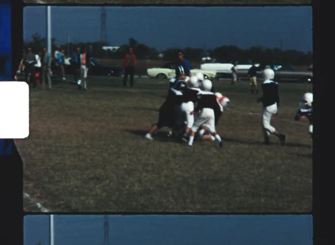 People watch young people play football on a field near a road lined with parked cars