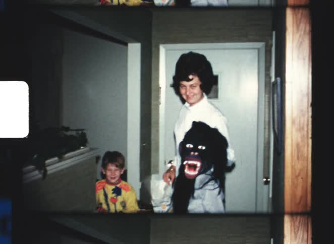 Woman in a home hallway beside two children one of them wearing a gorilla mask