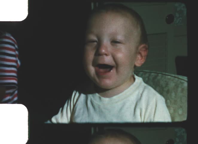 A toddler sitting on a chair laughing excitedly inside a room next to another kid
