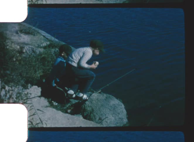Two young boys sit on a large rock at the edge of a dark river holding fishing rods captured in a vintage 8mm film look