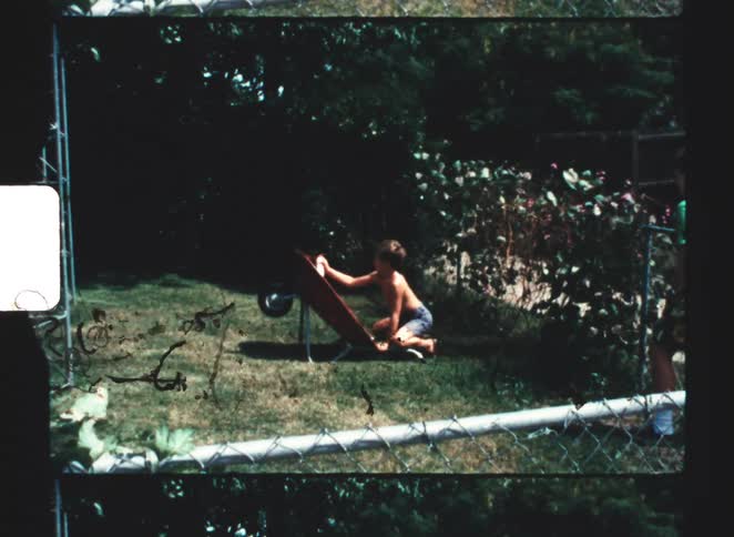 A shirtless kid spraypainting a wheelbarrow on a grassy lawn while another kid walks toward him