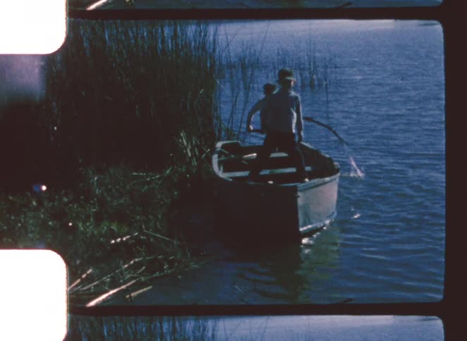 Two young boys in shorts and shirts stand in a small rowboat near the edge of a lake or river with tall reeds captured in a vintage 8mm film look