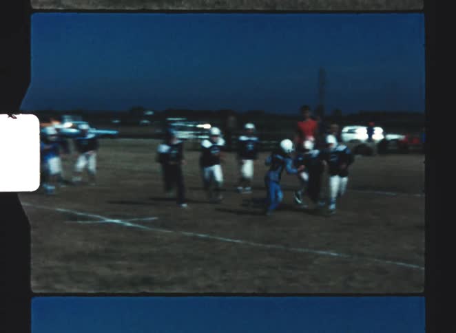 Youth football players wearing dark blue and white uniforms rush into a pile at the start of a play on a grassy field at dusk with several coaches and adults watching the action near the sideline