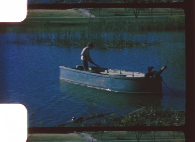 Two people are launching a small dark boat from a grassy shore into a body of water with tall reeds