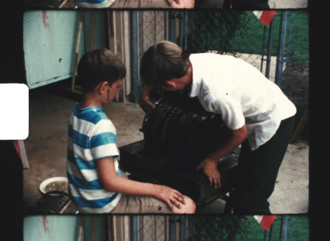 Two boys one in a striped shirt and the other in white are intently examining and adjusting a cash registerlike machine on a small cart outdoors