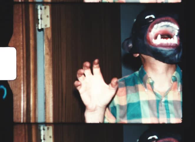 A man wears a horrifying black monkey mask with an enormous red and white mouth while raising his hand and shaking his fist indoors
