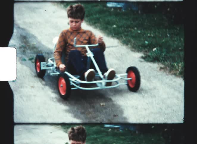 A young man with brown hair wears a plaid shirt and dark pants while riding a pedal powered go kart down a slight incline on a paved driveway captured in a vintage 8mm film look