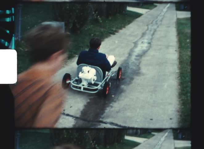 A father rides a pedal car as his son runs alongside in a quiet neighborhood alley