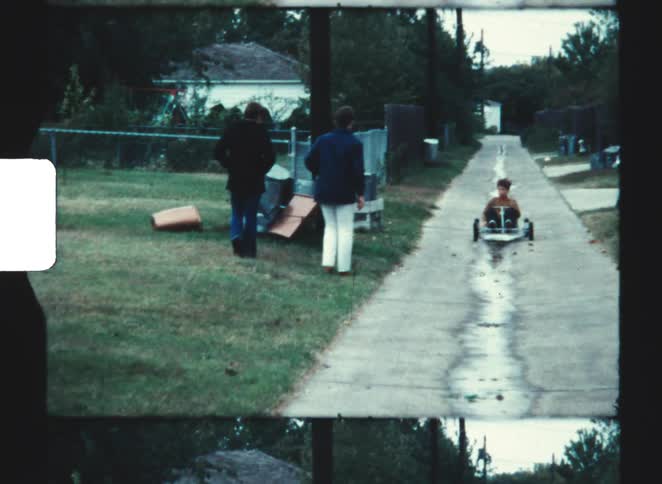 In a grainy home video a young boy slowly rolls down a long neighborhood driveway in a gokart as two adults watch from the lawn