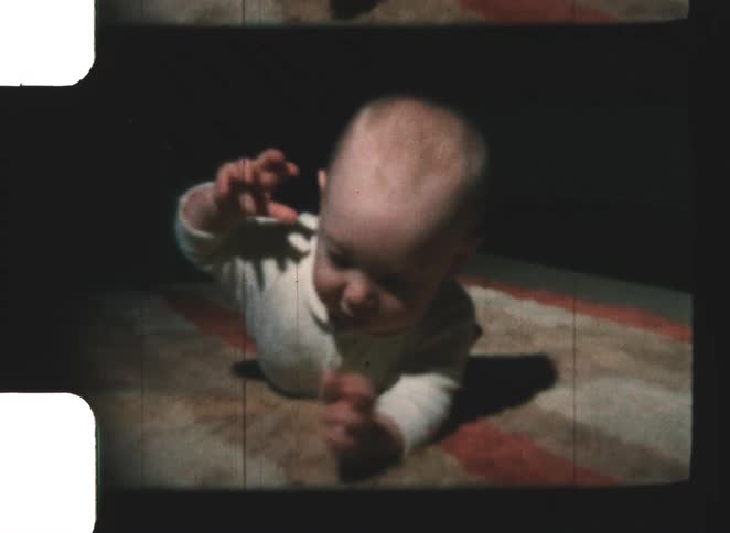 A bald baby wearing a white onesie lies on its stomach on a patterned rug looking directly at the camera while attempting to crawl smiling and lifting an arm