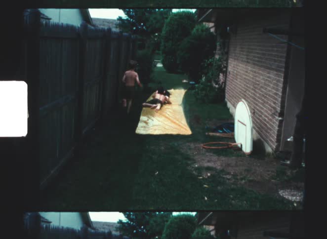 Two shirtless children are playing on a yellow slip n slide set up as a third runs past in a narrow backyard between a wooden fence and a brick wall